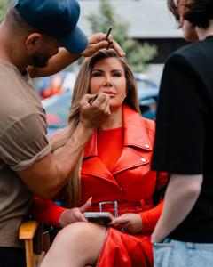 Adriana Gallardo in a bright red leather outfit sits confidently in a makeup chair while a makeup artist applies lip liner and another person observes. She holds her phone in one hand and looks poised, with long styled hair and a calm, focused expression.