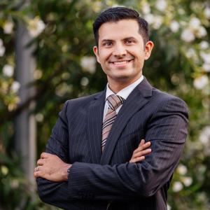 Dr Vivek Eranki, smiling in a suit with arms crossed, standing outdoors in front of greenery.