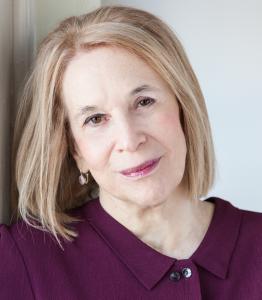 Sue William Silverman, award-winning author and memoirist, smiling gently while leaning against a wall. She has shoulder-length blonde hair, wears a plum-colored blouse, and exudes a warm and thoughtful expression.