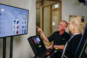A rehabilitation professional shows a patient the gaitACTIVE system on the screen. The professional is a middle aged white man and the patient is an older lady with blond hair.