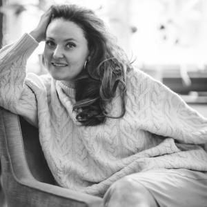 Charlotte Osler seated on a sofa in soft light, smiling warmly  representing her gentle, human-centered approach to aesthetic medicine.