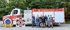 A group of seven people poses together in front of a fire truck labeled "Engine 602" in a forested area, showcasing community support.