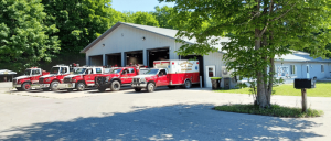A fleet of red emergency vehicles, including an ambulance, parked in front of a large metal fire station surrounded by greenery.