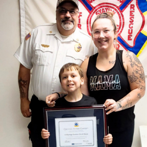 A family poses together, with a young boy holding a certification award, wearing casual attire in a celebratory setting.