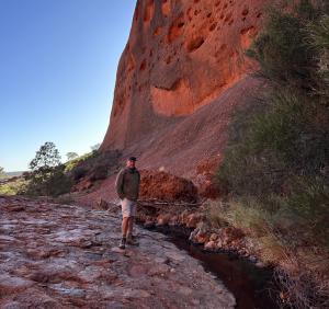 Paul Rushworth-Brown stands beside a stream at the base of Uluru, surrounded by red rock and native bushland, symbolising the novel’s deep connection to Country and Indigenous history.