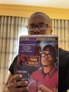 Kevin Davis, wearing glasses and a black shirt, holds up the debut issue of Kevysie International. The magazine cover features the same stylized artwork and highlighted interviews as the 3D version, with a focus on community, creativity, and emotional res