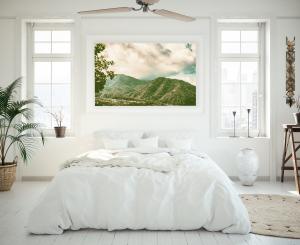 A bright, serene bedroom with a minimalist, natural aesthetic. The room features a large, plush white bed with soft linens and pillows, centered beneath a large landscape photograph of green mountains under a partly cloudy sky. The walls, floors, and furn