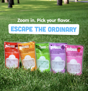 Red, orange, green, purple, and pink Jubes packs arranged upright on grass with the caption “Zoom in. Pick your flavor. Escape the Ordinary."