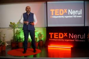 Dr. Mohana Rao Patibandla standing on a red circular carpet on stage at the TEDx Nerul event, wearing a blue vest and white shirt, holding a microphone. The backdrop features the TEDx Nerul logo and the text "X=independently organized TED event" on a scre