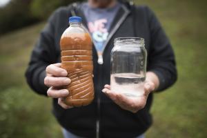 A close up shot of a woman's hands. In one hand is a water bottle filled with brown liquid. In the other hand is a clear mason jar filled with clean water.