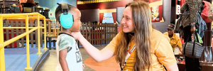 A smiling staff member, wearing a yellow shirt, adjusts headphones on a young child in a colorful indoor play area.