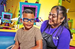 A boy with a brown shirt and blue glasses sitting in a kid-sized salon chair with his barber admiring his new hair style