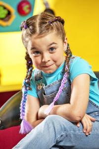 A girl in overalls and Dutch braids sits in a kid-sized chair at a salon with her arms crossed.