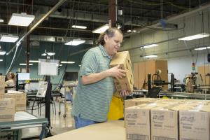 Smiling man in a striped shirt holding boxes in the Austin Lighthouse warehouse, surrounded by packages ready for shipping.