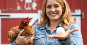 As food prices rise and sustainability becomes a priority, families across the U Women holding a chicken and fresh egg with red bard in background.