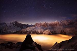 A cloud drapes over a valley, glowing orange from the city lights below, with mountain peaks protruding through the mist and a starry night sky above.