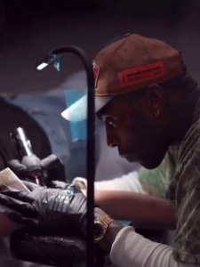 Tattoo artist Derrick Johnson concentrating while tattooing a client, wearing gloves and a cap under focused lighting.