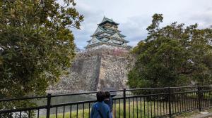 Two boys standing near the moat, looking at Osaka Castle’s stone walls and historic tower in Osaka, Japan, during a family trip.