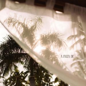 Sombras de palmeras proyectadas sobre una cortina blanca con luz cálida al atardecer. En la esquina inferior derecha aparece el título del sencillo ‘A Falta De’ de ISA una cantautora y productora dominicana.