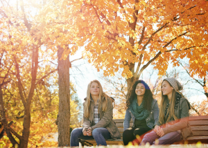 Three young women sitting together on a wooden bench in a park with vibrant autumn foliage, dressed in casual fall clothing and appearing relaxed and happy