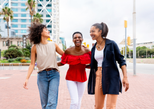 Three young women of diverse backgrounds laughing together outdoors in an urban plaza setting with modern buildings and palm trees in the background