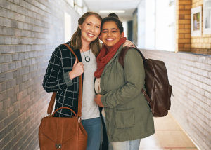 Two diverse female college students smiling and embracing in a brick-lined school hallway, one wearing a plaid jacket and carrying a leather bag, the other in an olive jacket with a backpack