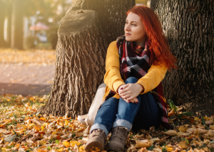 A woman with red hair in a yellow sweater and plaid scarf sitting peacefully against a tree trunk surrounded by autumn leaves, looking contemplatively into the distance