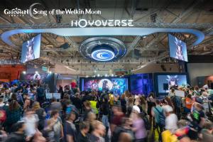 An aerial view of the crowded Genshin Impact booth at Gamescom 2025. Large screens show character art and gameplay, with a dense crowd of fans and visitors below. Motion blur highlights the event's high energy.