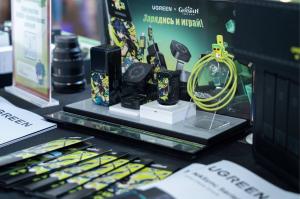 A close-up view of a display featuring UGREEN x Genshin Impact co-branded products at a gamescom booth. On a tiered platform, several dark-colored power banks and a charging cube are visible with bright green character art from the game. A coiled, yellow-