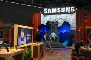 A detailed view of the Genshin Impact trial area at the Samsung booth during gamescom. The scene is dominated by a themed throne, built to resemble an ancient stone seat, where a fan sits for a photo. In the foreground, Samsung devices on wooden stands di