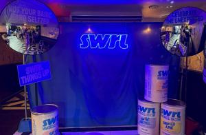 Photo from SWRL's NYC launch party showing a blue neon SWRL sign, oversized branded cans, and two photo mirrors with event slogans.