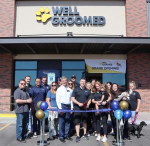 Well Groomed team members gather outside a salon for a grand opening celebration. A ribbon is stretched across the entrance with balloons, and a banner above the door reads “Well Groomed Grand Opening!” Several team members hold dogs, highlighting the bra