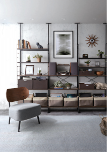 A modern living room featuring a large, black, floor-to-ceiling modular shelving unit filled with books, plants, and a record player, with a chic grey and wood chair in the foreground.