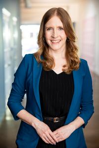 Cheryl Platz is a smiling blonde professional woman with a blue blazer and a black pinstripe matched tank top and pantsuit set with belt. Her hands are folded in front of her body and she smiles softly to the camera.
