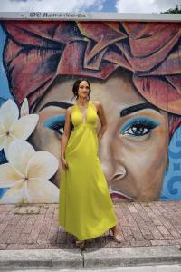 Camille Thomas, Miss Universe Curaçao 2025, stands elegantly in a full-length, vibrant yellow halter dress while posing in front of a large, colorful street art mural of a woman's face.