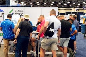 A group of people gather around the Brelly and Building Experts Institute booth at a conference, watching a live demo on a laptop.