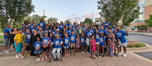 Group of students wearing royal blue custom spirit week t-shirts, smiling and raising arms outside a school building.