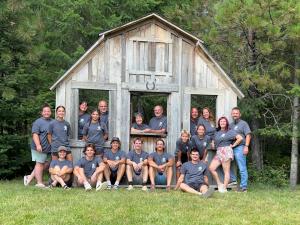 Group of teens and adults in gray custom t-shirts posing by a rustic wooden cabin in a camp setting.