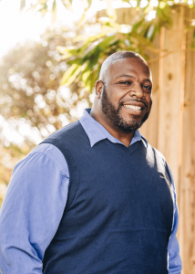Yeates Family Consulting Expands Comprehensive Mental Healthcare Access with Digital Innovation 16 Edward Yeates, a smiling Black man wearing a blue sweater vest over a light blue dress shirt, stands confidently outdoors with warm sunlight and greenery in the background.