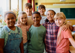 A diverse group of seven elementary school-aged children smiling and embracing each other in a bright classroom environment.