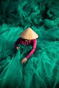 A top-view image of a Vietnamese woman in a conical hat, dressed in magenta, delicately handling large green fishing nets in a fine art photograph by Réhahn.
