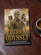 Paperback copy of Outback Odyssey by Paul Rushworth-Brown resting on a wooden table beside a cup of tea, symbolizing reflection and quiet conversation amidst social unrest.