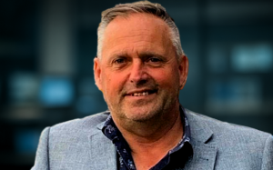 Middle-aged man in a grey blazer standing in front of a blurred newsroom background, symbolizing his engagement with media and contemporary cultural discussions.