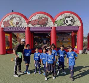 A group of young soccer players in matching blue “Adriana’s” jerseys stands with their coach on a turf field in front of inflatable game booths labeled “Fast Ball,” “Football Toss,” and “Penalty Kick.” The kids are smiling and posing together in the sunli