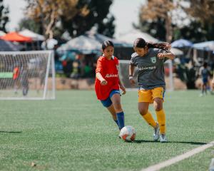 Two girls in soccer jerseys, one in red and one in gray with yellow shorts, battling for possession of the ball on a turf field.