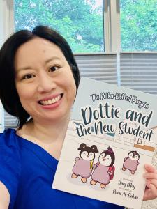 Asian woman with short bob and smiling expression holds up copy of hardcover book Dottie and the New Student, showing 3 baby penguins wearing backpacks on an iceberg.