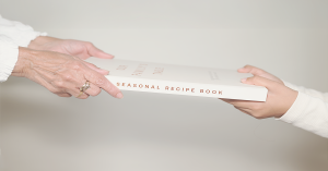 Glimmery Moment Hands passing the Our Family’s Table recipe journal between generations, symbolizing offline gifting and shared heritage