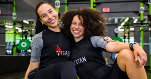 Two women in Titan Body EMS training suits smiling and posing at the gym with fitness equipment in the background.