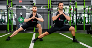 Two men wearing Titan Body EMS training suits doing synchronized side lunges on turf in a gym with weight racks in the background.