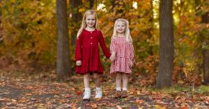 Two smiling girls in Mabel + Honey fall dresses hold hands on a path covered in vibrant autumn leaves.